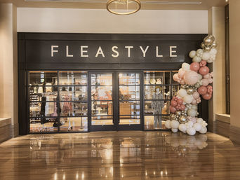 Stylish indoor shopping mall boutique storefront with glass doors, clothing displays and mannequins, festive pink, white and gold balloon garland at the entrance and warm lights reflecting on the polished floor.