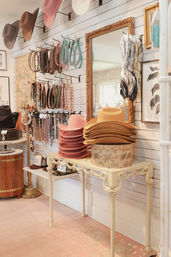 Bright boutique interior with stacked pink and tan felt cowboy hats on a vintage console table, wall racks of belts, necklaces and colorful scarves hung beside an ornate mirror — stylish accessories display.
