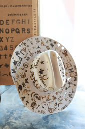 Cream hand-stamped cowboy hat covered in playful burned western symbols — horseshoes, stars, cacti and 'YEEHAW'/'TEXAS' lettering — leaning against an alphabet stencil board on a blue marbled surface.