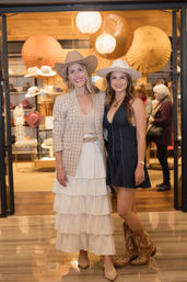 Two women wearing wide-brimmed cowgirl hats and stylish outfits — a tiered white skirt and a denim dress with embroidered cowboy boots — posing at a boutique hat shop entrance with hat displays and warm lighting.