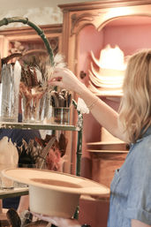 Person selecting decorative feathers from glass jars to trim a tan felt hat in a boutique millinery studio with stacked hats in the background