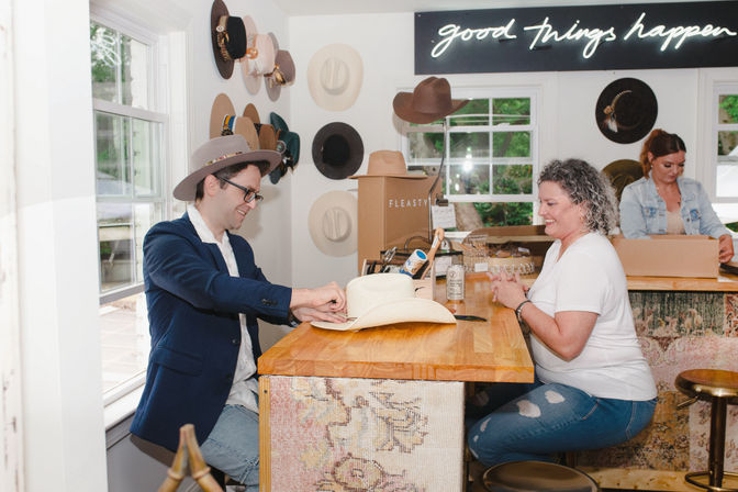 Inside a cozy hat boutique, two people at a wooden counter try on and admire cowboy hats beneath a neon “good things happen” sign, with wall-mounted hat displays and a staff member packing orders in the background.
