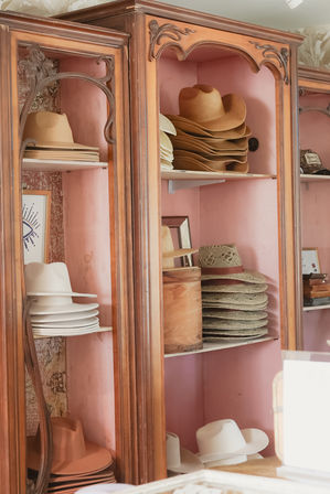 Stacks of tan, white and woven straw cowboy hats displayed on vintage wooden shelves with a pink-painted backing.