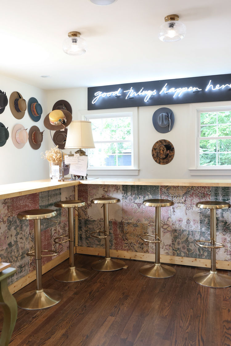 Cozy bright cafe-style counter with five brass bar stools, patterned bar front, wall of decorative hats and neon sign reading 'good things happen here' above two windows