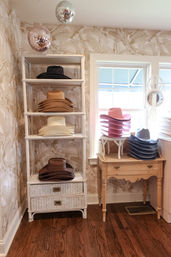 Sunlit boutique corner with stacked cowboy hats on white wicker shelves and a small wooden table by a window — pink, gray, tan and brown hats, floral wallpaper and hanging disco balls.