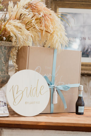 Wedding gift display: bridal gift box tied with pale blue ribbon, pampas grass arrangement and mini champagne bottle on a rustic wooden table