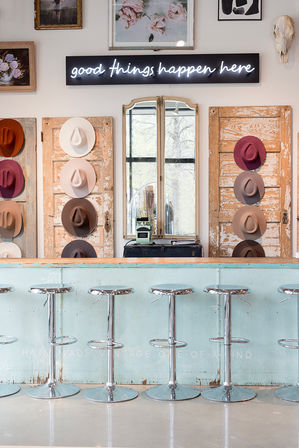 Cheerful boutique interior with row of chrome bar stools at a distressed turquoise counter, vintage hat display on reclaimed doors, central mirror and neon sign reading 'good things happen here'.