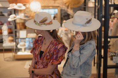 Two women trying on white wide-brim feathered hats in a stylish boutique hat shop, one wearing a colorful dress and the other a denim jacket.