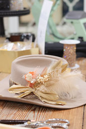 Beige felt wide-brim hat adorned with cream feathers, dried flowers and a golden ribbon, resting on a wooden worktable in a hat-making workshop