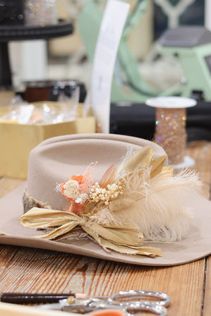 Beige felt wide-brim hat adorned with cream feathers, dried flowers and a golden ribbon, resting on a wooden worktable in a hat-making workshop