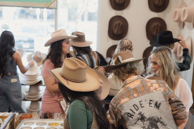 Women browsing and trying on tan and black cowboy hats in a western hat boutique, with a wall display of hats and an accessory counter visible.