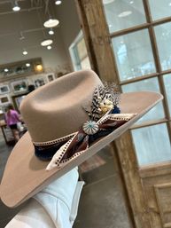 Tan wide-brim felt western hat decorated with a brown bandana-style ribbon, spotted feathers and a turquoise concho, held against a rustic boutique interior