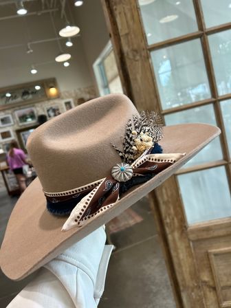 Tan wide-brim felt western hat decorated with a brown bandana-style ribbon, spotted feathers and a turquoise concho, held against a rustic boutique interior