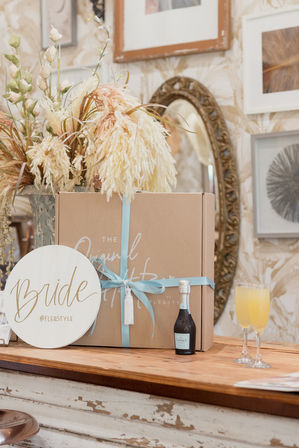 Bridal shower welcome box — kraft gift box tied with light blue ribbon on a rustic wooden counter, flanked by a dried floral arrangement, a small sparkling wine bottle and two mimosas, with a vintage mirror and framed art in the background.
