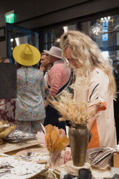 Blonde shopper examining jewelry on a wooden table with dried floral arrangements at an indoor boutique pop-up market