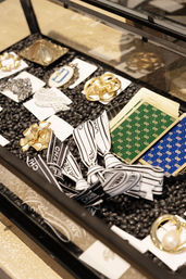 Boutique glass display of luxury accessories: gold and pearl brooches, decorative pins, striped logo ribbons and patterned cards arranged on a black pebble tray.