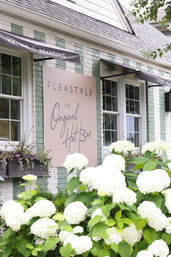 Charming pastel-painted brick hat shop storefront with a large blush sign for a hat bar, dark awnings, window boxes, and abundant white hydrangea blooms in the foreground.