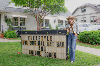 Woman in a cowboy hat and denim leaning on a vintage marquee sign promoting a hat bar and unique goods outside a white-brick storefront with trees and parked cars