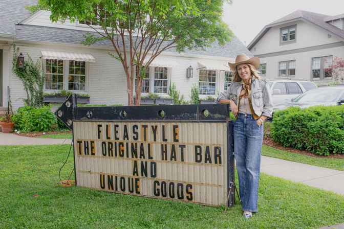 Woman in a cowboy hat and denim leaning on a vintage marquee sign promoting a hat bar and unique goods outside a white-brick storefront with trees and parked cars