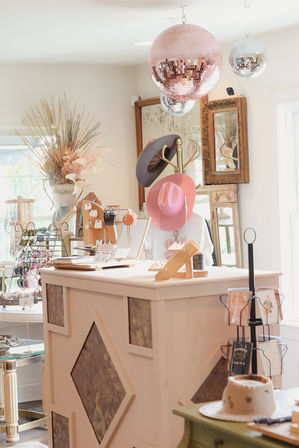 Bright pastel boutique interior with pink disco ball, hat rack, jewelry and sunglasses displays on a wooden checkout counter, dried floral arrangement and vintage mirror