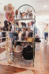 Sunlit hat boutique interior with a vintage green rack displaying stacked cowboy hats and jars of feather trims for customizing hats.
