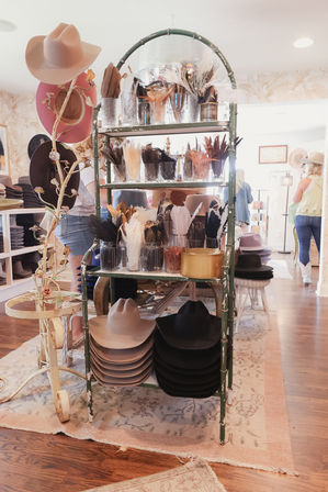 Sunlit hat boutique interior with a vintage green rack displaying stacked cowboy hats and jars of feather trims for customizing hats.