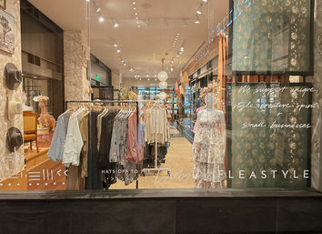 Boutique clothing store window display with racks of women's dresses, blouses and hats, mannequins in floral dresses and warm, inviting lighting