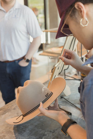 Close-up of a milliner steaming and shaping a tan felt cowboy hat at a boutique hat shop counter, hands adjusting the brim while a customer waits in the background.