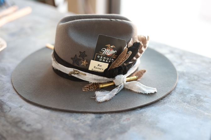 Gray wide-brim felt fedora on a marble countertop, decorated with layered ribbons, star pins, feathers, a tucked black tag and a brass bullet accent.