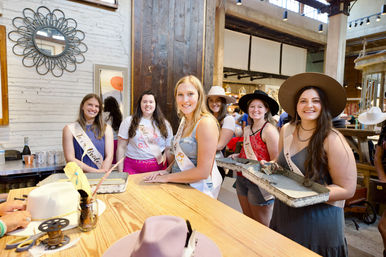 Six women wearing sashes and wide‑brim hats smiling at a rustic hat‑making workshop, gathered around a wooden table with hat blocks, trims and trays — playful bachelorette party in a boutique studio.