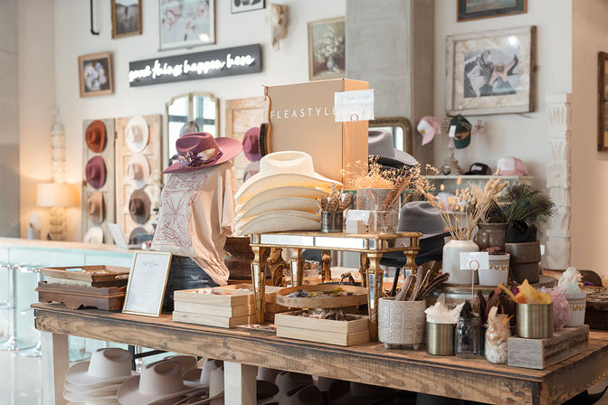 Boho boutique display of wide-brim hats stacked on a rustic wooden table, a purple felt hat on a mannequin, dried floral arrangements, and assorted accessories and gift boxes in a bright retail shop.