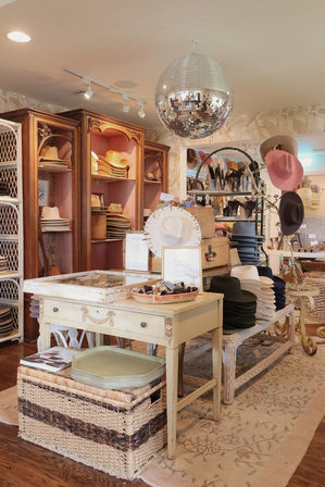 Boutique hat shop interior with stacked straw and felt hats on wooden cabinets and wicker displays, vintage cream display table and mirrored disco ball overhead