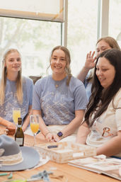Smiling women in matching light-blue celebration tees gathered around a wooden table with mimosas and a bottle in a sunlit indoor café, cheerful bridal-brunch vibe.