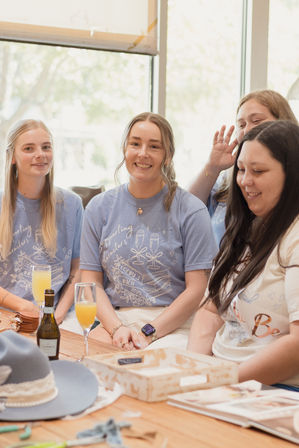Smiling women in matching light-blue celebration tees gathered around a wooden table with mimosas and a bottle in a sunlit indoor café, cheerful bridal-brunch vibe.
