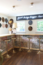 Bright boutique hat shop interior with hats displayed on walls, light wood bar counter with five brass swivel stools, patterned fabric bar front, neon sign reading 'good things happen here', and windows letting in natural light.