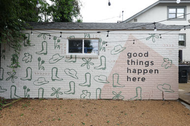 Brick courtyard wall mural with playful green hat and boot doodles, pink triangular accent and bold black text reading “good things happen here,” string lights overhead and gravel ground.