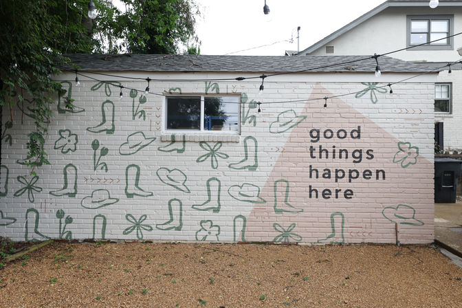 Brick courtyard wall mural with playful green hat and boot doodles, pink triangular accent and bold black text reading “good things happen here,” string lights overhead and gravel ground.