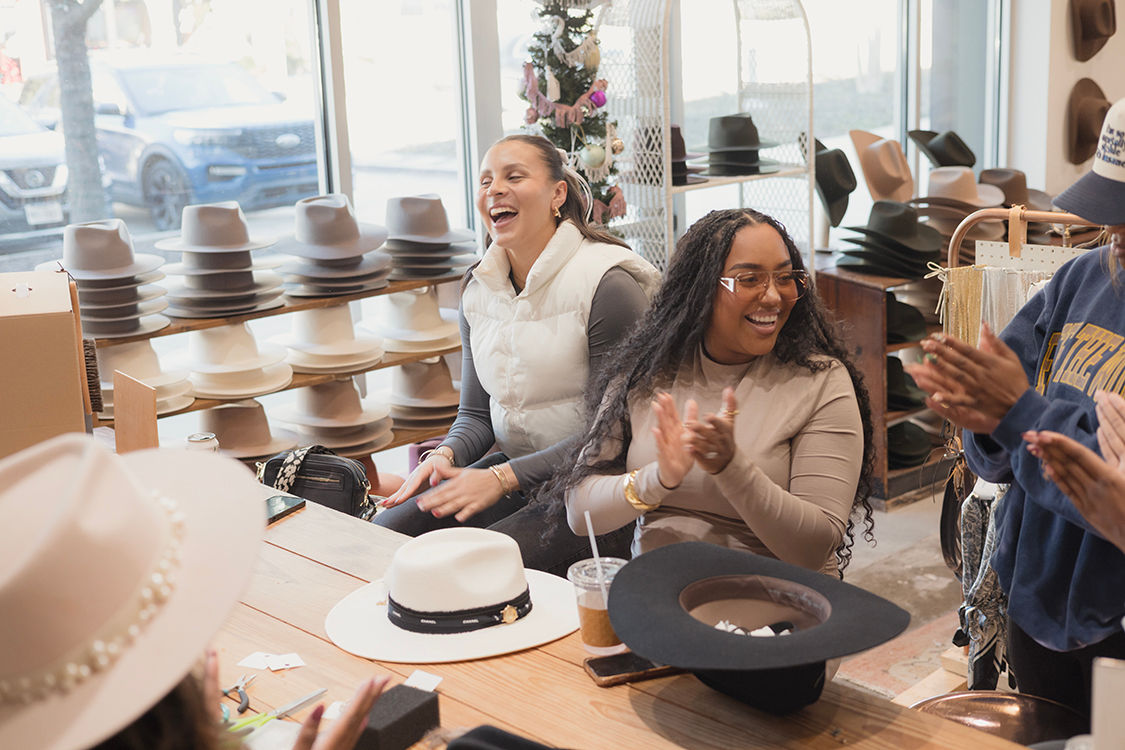 Three women laughing and clapping in a sunlit hat boutique, trying on stylish wide-brim and fedora hats around a wooden table.