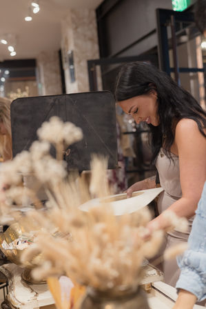 Smiling woman with long dark hair shopping for a beige wide‑brim hat at a chic boutique counter, surrounded by dried flower arrangements and decorative display bowls