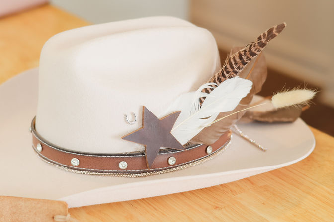 Light beige western-style felt hat on a wooden table, cowboy-chic with a brown leather band studded with rhinestones, a wooden star pin and mixed decorative feathers.