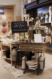 Boutique hat bar station with a sign, trays of feathers and trims, stacked hats on shelves and a vintage suitcase display in a cozy accessory shop