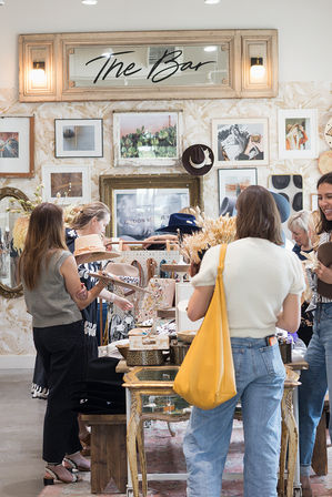 Women browsing hats, jewelry and accessories at a bright, boho-style boutique display table with framed art and a decorative mirror gallery wall.