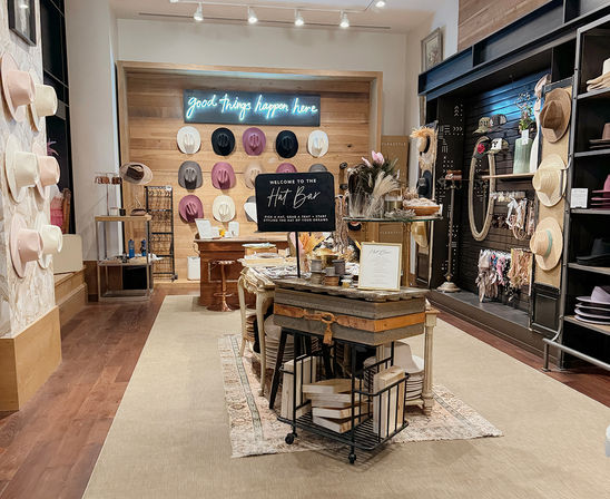 Stylish hat boutique interior with wall-mounted fedoras and straw hats, central 'Hat Bar' table of trims and accessories, neon “good things happen here” sign and warm wood floors