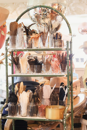 Decorative feather trims in glass jars on a chipped green metal display shelf in a hat boutique, with stacked felt hats and a disco ball overhead.