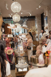Busy boutique hat shop interior with shoppers browsing wide-brim hats and accessories beneath sparkling crystal globe chandeliers.