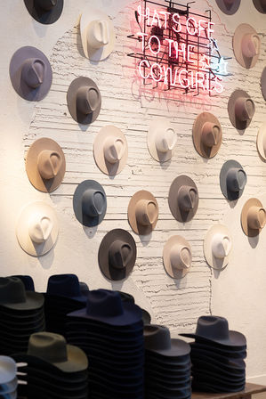 Retail display of pastel and neutral felt cowboy hats mounted on a textured white wall with stacked hat piles in the foreground and a playful pink neon sign reading 'HATS OFF TO THE COWGIRLS'.