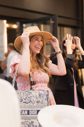 Smiling woman in a floral dress trying on a tan wide‑brim hat in a downtown boutique, holding a patterned canvas tote bag — a candid stylish shopping moment.