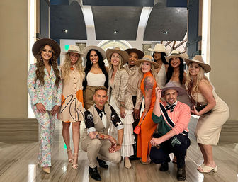 Smiling group of eleven people in stylish outfits and wide‑brim hats posing for a group photo in an upscale hotel lobby with arched ceiling, marble floors, and warm lighting.