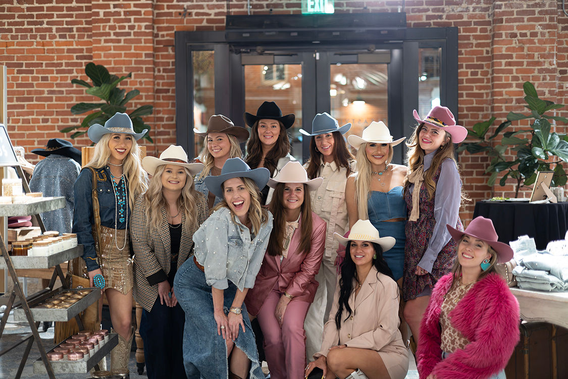 Smiling group of women wearing colorful cowboy hats and Western-style outfits posing inside a brick-walled urban loft boutique at a cowgirl fashion pop-up event.