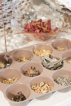 Close-up of a compartment organizer on a DIY jewelry workspace holding sorted gold, silver and rose-gold findings — jump rings, clasps, small keys and connectors — with a decorative glass bowl of colorful craft pieces in the background.